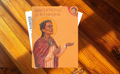 A religious art print or icon card titled 'Saint Catherine of Alexandria' featuring an illustration of the saint in a red robe and gold halo, placed on a wooden surface.